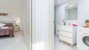 A modern bedroom with a bed, bedside table, and a nightstand. A bathroom with a sink, mirror, and a washing machine is visible.
