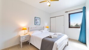Ein Hotelzimmer mit einem Bett, zwei Nachttischen mit Lampen, einem Deckenventilator, einem Fenster mit Vorhängen und einer Tür.
