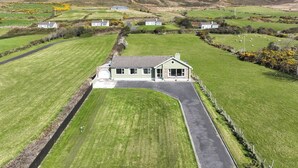 A large, single-story house with a paved driveway and a well-maintained lawn, surrounded by rolling hills and a mountain in the background.