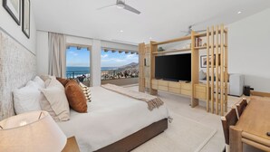 A modern bedroom with a large bed, a TV, and a balcony with a view of the sea.