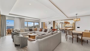 A spacious living area with a large sofa, a dining table, and chairs. There are large windows offering a view of the outdoors.
