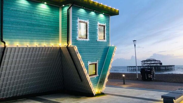 A teal building with a slanted roof and a small window, located near a beach with a pier in the background. - Mill Cottage | By My Getaways (Brighton)