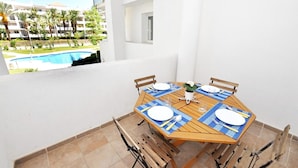 Mesa de comedor preparada para cuatro personas, con individuales azules, sillas de madera y vista a una piscina y edificios al fondo.