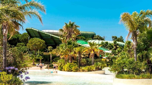 Un parque acuático con un tobogán verde grande, palmeras y una piscina cristalina.