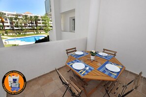 A dining table set for four with blue placemats, white plates, and cutlery, positioned on a balcony with a view of a swimming pool and palm trees. - Apartment in Roses near Sandy Beach (Rosas / Roses)