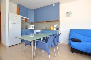 A compact kitchen with a refrigerator, microwave, and coffee maker, a glass dining table with chairs, and a blue sofa.