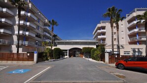 A multi-story hotel entrance with a clear sky, palm trees, and a parking area with a handicapped parking space. - Apartment in Roses with large terrace (Rosas / Roses)