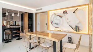 Un espace repas moderne avec une table en bois et des chaises à structure métallique, une cuisine en arrière-plan et un luminaire fixé au mur.