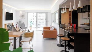 Une cuisine moderne avec des luminaires suspendus blancs, un coin repas avec une table et des chaises en bois, et une grande fenêtre offrant une vue sur la ville.