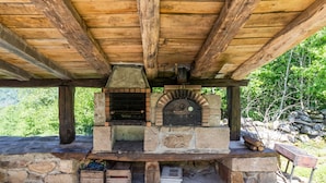Une cuisine extérieure en pierre, dotée d’un four en briques et d’un barbecue au bois, le tout intégré dans un décor naturel composé d’arbres et de rochers.