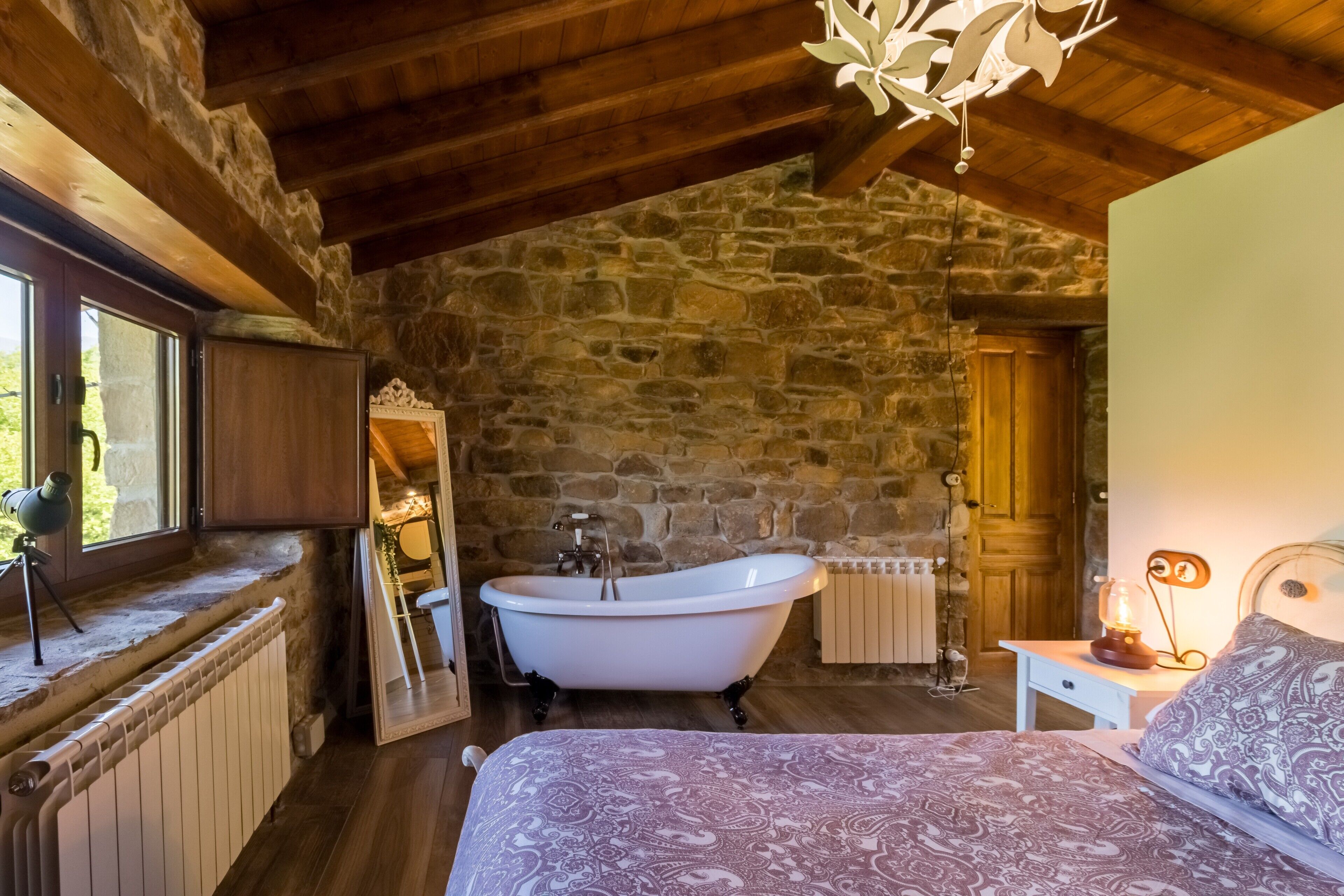 Un dormitorio rústico con pared de piedra, techo de madera, una cama grande, una bañera de patas de león y un telescopio cerca de la ventana.