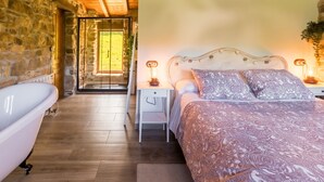 Une chambre avec un mur en pierre, un plancher en bois, une baignoire blanche, un lit avec une literie à motifs et une table de chevet avec une lampe.