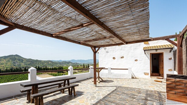 A covered outdoor area with stone flooring, wooden benches, and a view of distant hills.