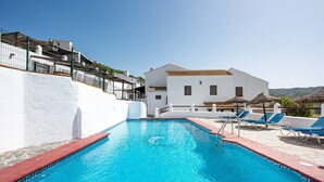 A swimming pool with a red border, a white building with a tiled roof, and blue lounge chairs.