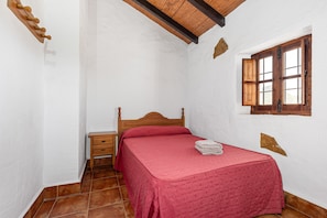 A bedroom with a wooden bed, a nightstand, a window with wooden shutters, and a white wall with a small stone decoration.