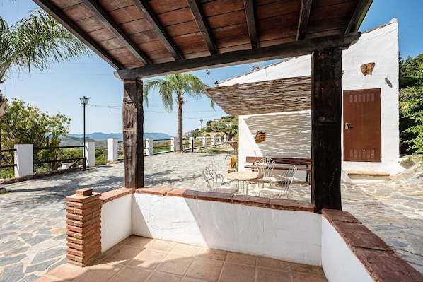 A covered outdoor seating area with stone flooring, a wooden pergola, and a white building with a brown door.