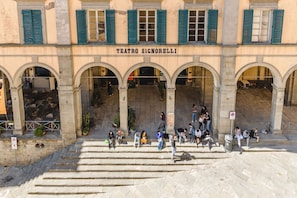 A building with 'TEATRO SIGNORELLI' signage, multiple arches, and people sitting and walking in front.