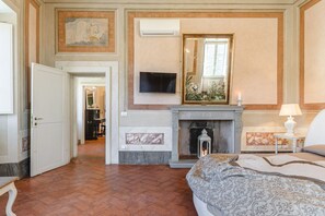 A hotel room with a fireplace, a television mounted on the wall, a bed with patterned bedding, and a hallway leading to another room. - Appartamento Centrale - Overlooking the charming Piazza Signorelli (Cortona)