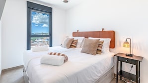 Ein Schlafzimmer mit einem großen Bett, Blick auf die Stadt durch ein Fenster, einem Nachttisch mit Lampe und einer Deckenleuchte.