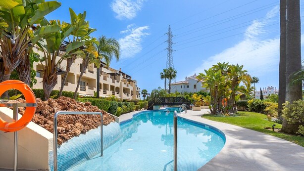 A swimming pool with a lifebuoy, surrounded by palm trees and a building in the background. - Stunning Views-Miraflores Apartment; Pass the Keys (Mijas)