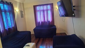 Une chambre avec deux lits recouverts d’un tissu sombre, un petit pouf blanc et un téléviseur à écran plat fixé au mur.