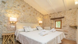 Un dormitorio con pared de piedra, una cama con ropa de cama blanca, dos lámparas de noche, una mesita de madera y una ventana con vistas.