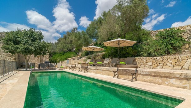 Una piscina con sillas de descanso y sombrillas, rodeada de muros de piedra y árboles.