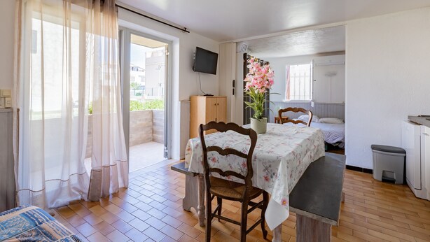 Een kamer met een houten vloer, een eettafel met stoelen, een aan de muur bevestigde televisie, een bed, een kleine prullenbak en een raam met transparante gordijnen.