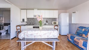 Een compacte keuken met witte kastjes, een eettafel met een tafelkleed met bloemenprint, een kamerplant en een blauwe gestreepte bank.
