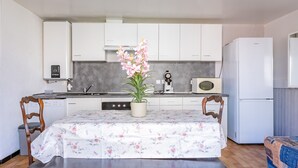 Een compacte keuken met witte kastjes, een magnetron, een fornuis en een koelkast. Er staat een tafel met een tafelkleed met bloemenprint en een potplant.
