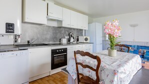 Een moderne keuken met witte kasten, een vaatwasser, een oven en een spoelbak. Er staat een eettafel met een tafelkleed met bloemen en een potplant.
