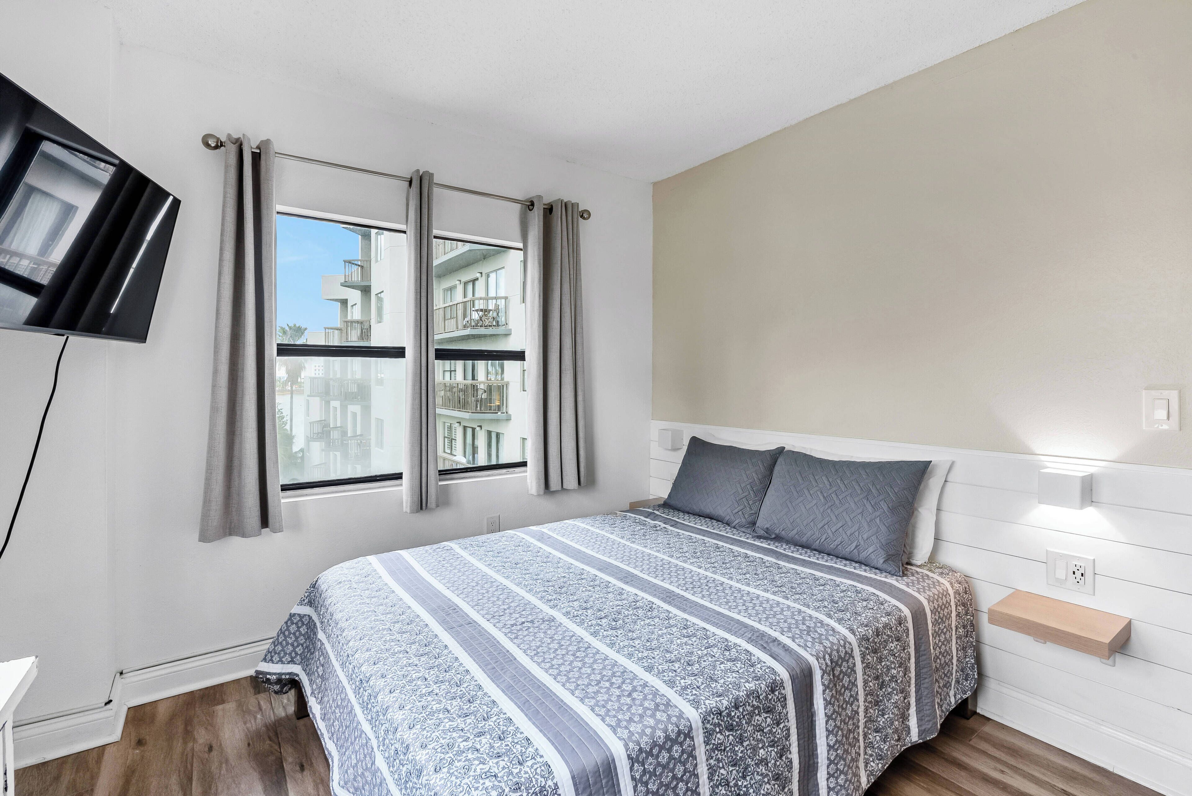 This cozy bedroom offers a comfortable bed with a stylish striped blanket, soft beige walls, and plenty of natural light from large windows.