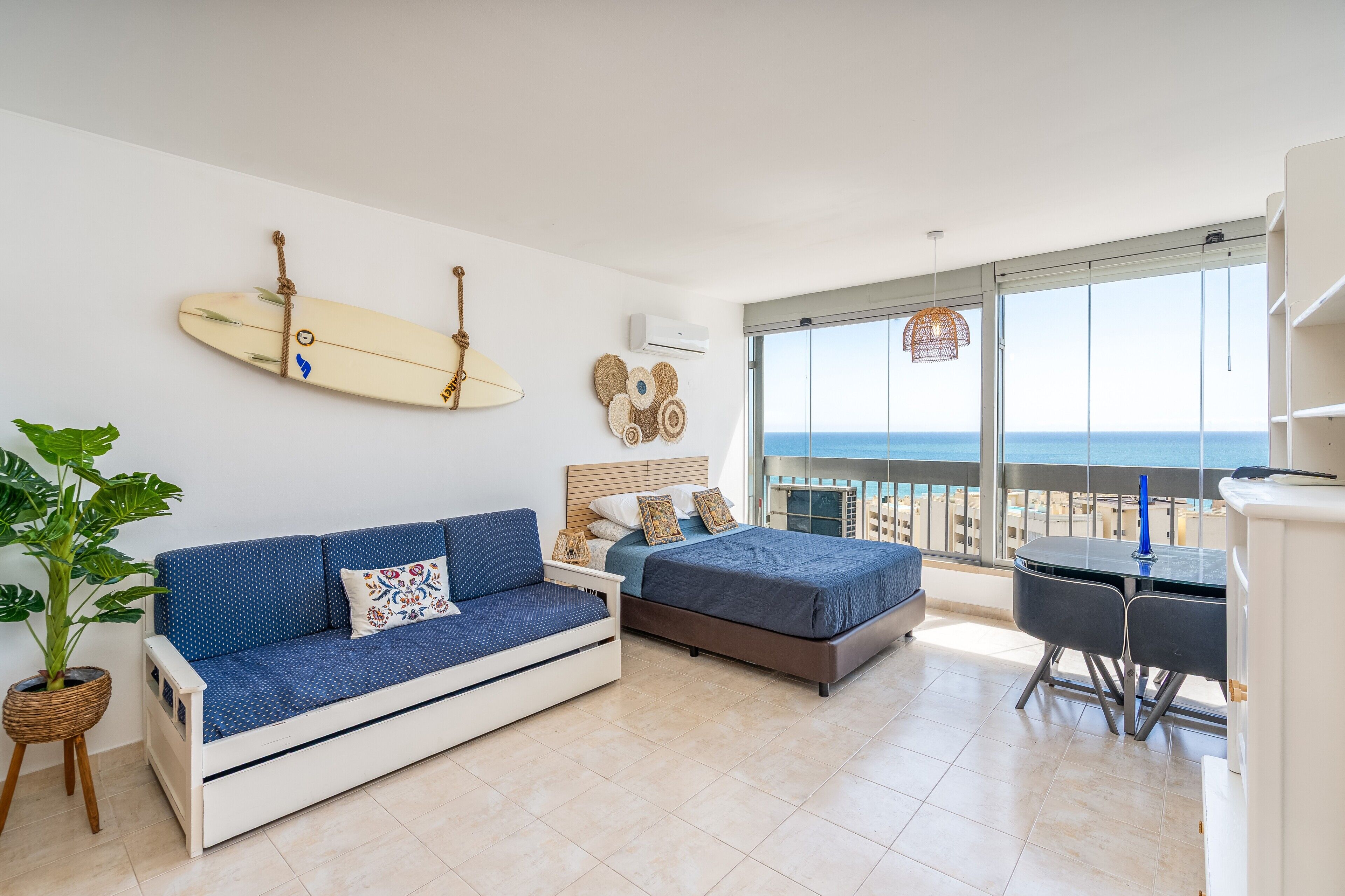 A bedroom with a bed, a sofa, a dining table, and a balcony with a view of the sea.