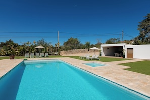 A rectangular swimming pool with a stone deck, surrounded by green grass and trees. There are lounge chairs and a covered patio area with a table and chairs.