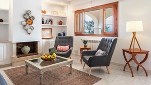 Un salon avec une cheminée, deux fauteuils, une table basse avec des fruits, une table d’appoint et une grande fenêtre donnant sur un beau paysage.