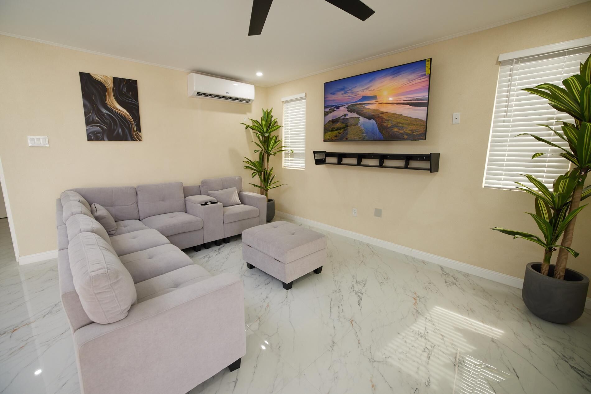 A living room with a sectional sofa, a small ottoman, a ceiling fan, a mounted air conditioner, a landscape painting, a shelf with decorative items, and potted plants.