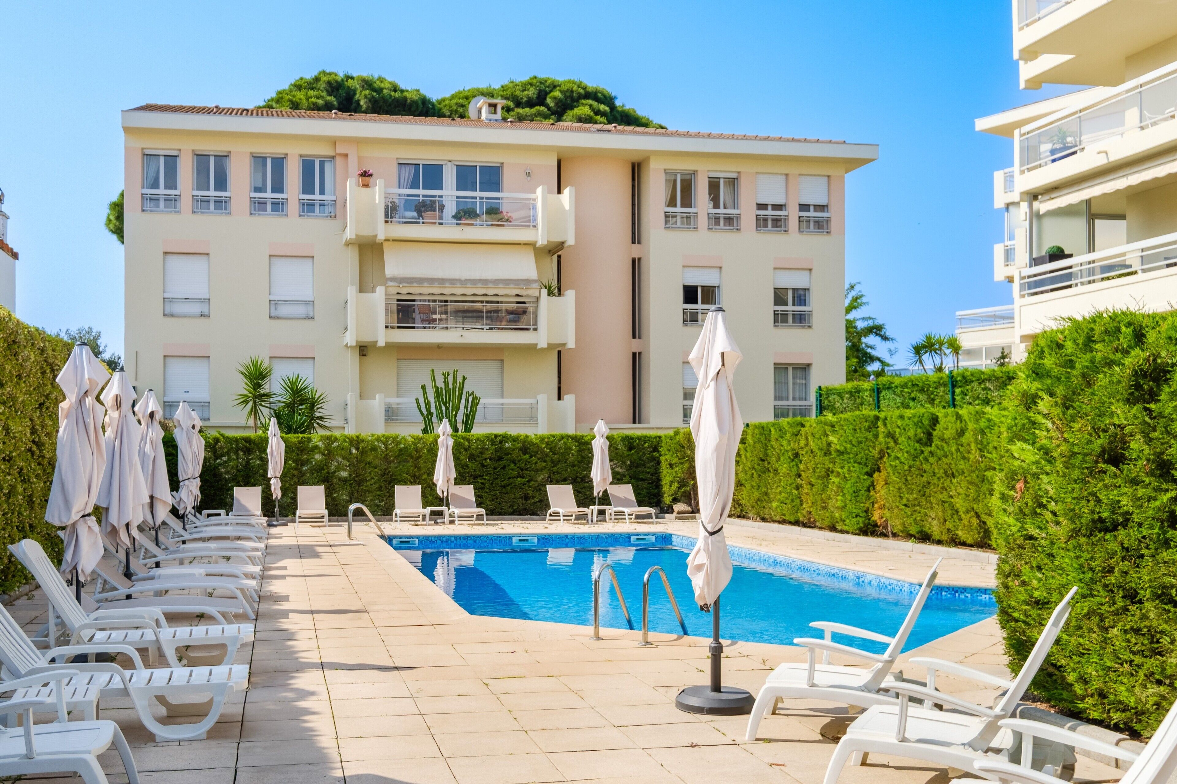 Un espace aménagé au bord de la piscine, avec des chaises longues et des parasols, une piscine et un bâtiment doté de balcons.