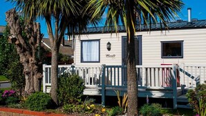A mobile home with a deck, surrounded by palm trees and a brick wall. - Introducing the ABI Vista, a 2-bedroom caravan designed to accommodate up to 6 guests. (Caister-on-Sea)