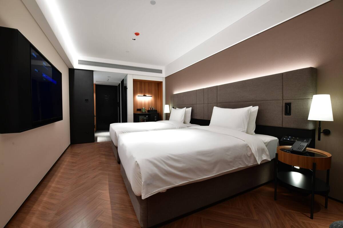 A modern hotel room with a large bed, a bedside table with a lamp, a flat-screen TV mounted on the wall, and a wooden floor.