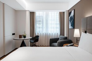 Habitación de hotel con una cama, un escritorio con silla, una mesita con una planta, una lámpara y un cuadro en la pared.