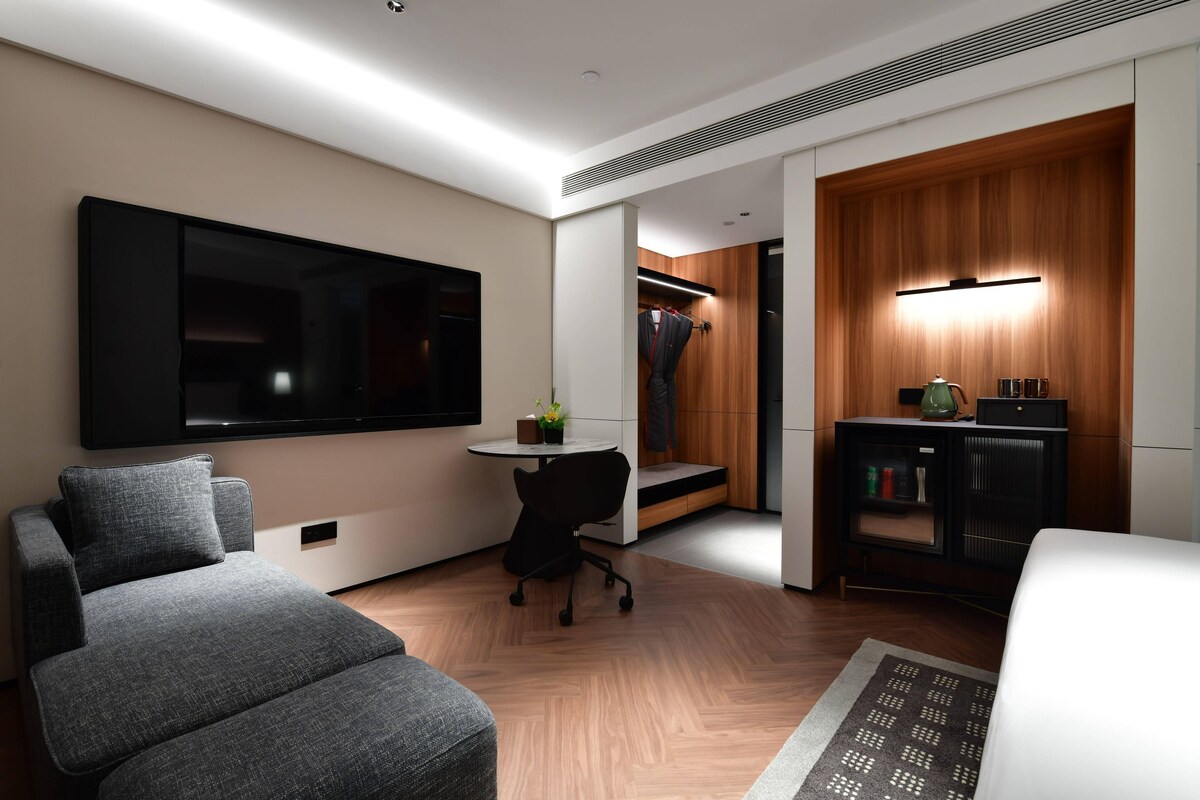 Une chambre d’hôtel moderne équipée d’un téléviseur à écran plat, d’un canapé gris, d’une petite table ronde avec une chaise, d’une armoire en bois et d’une bibliothèque.