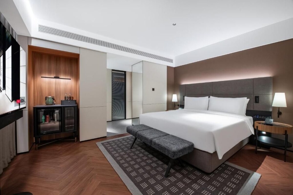 Ein modernes Hotelzimmer mit einem großen Bett, einer Bank, einem Nachttisch, einem Schrank und einem Spiegel.