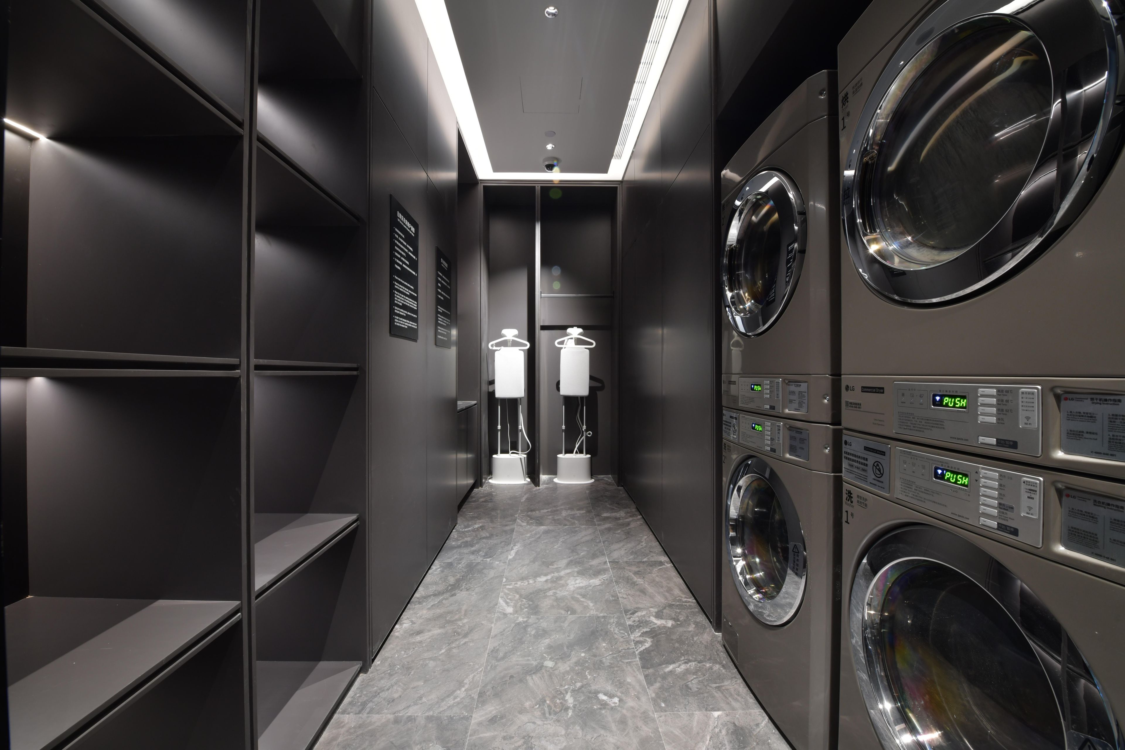 A modern laundry room with multiple washing machines and dryers, a tiled floor, and a narrow hallway.