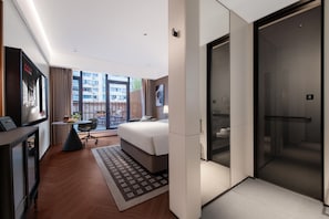 A modern hotel room with a bed, a desk, a chair, a TV, a balcony with a view of the city, and a sliding door to the bathroom.