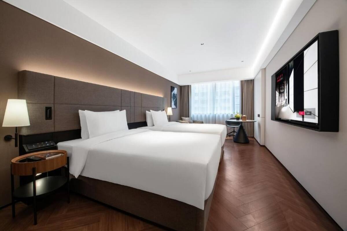 A hotel room with two beds, a desk, a chair, a lamp, and a large window.