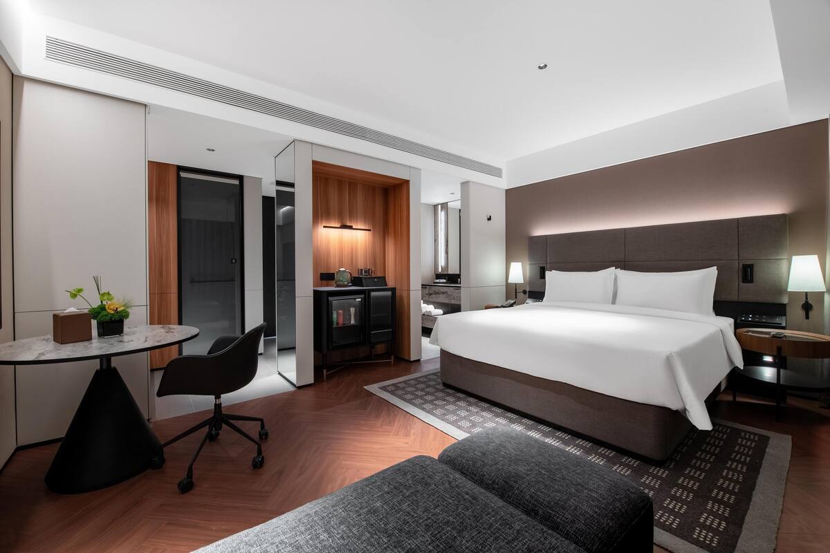 A modern hotel room with a large bed, a desk, and a kitchenette.