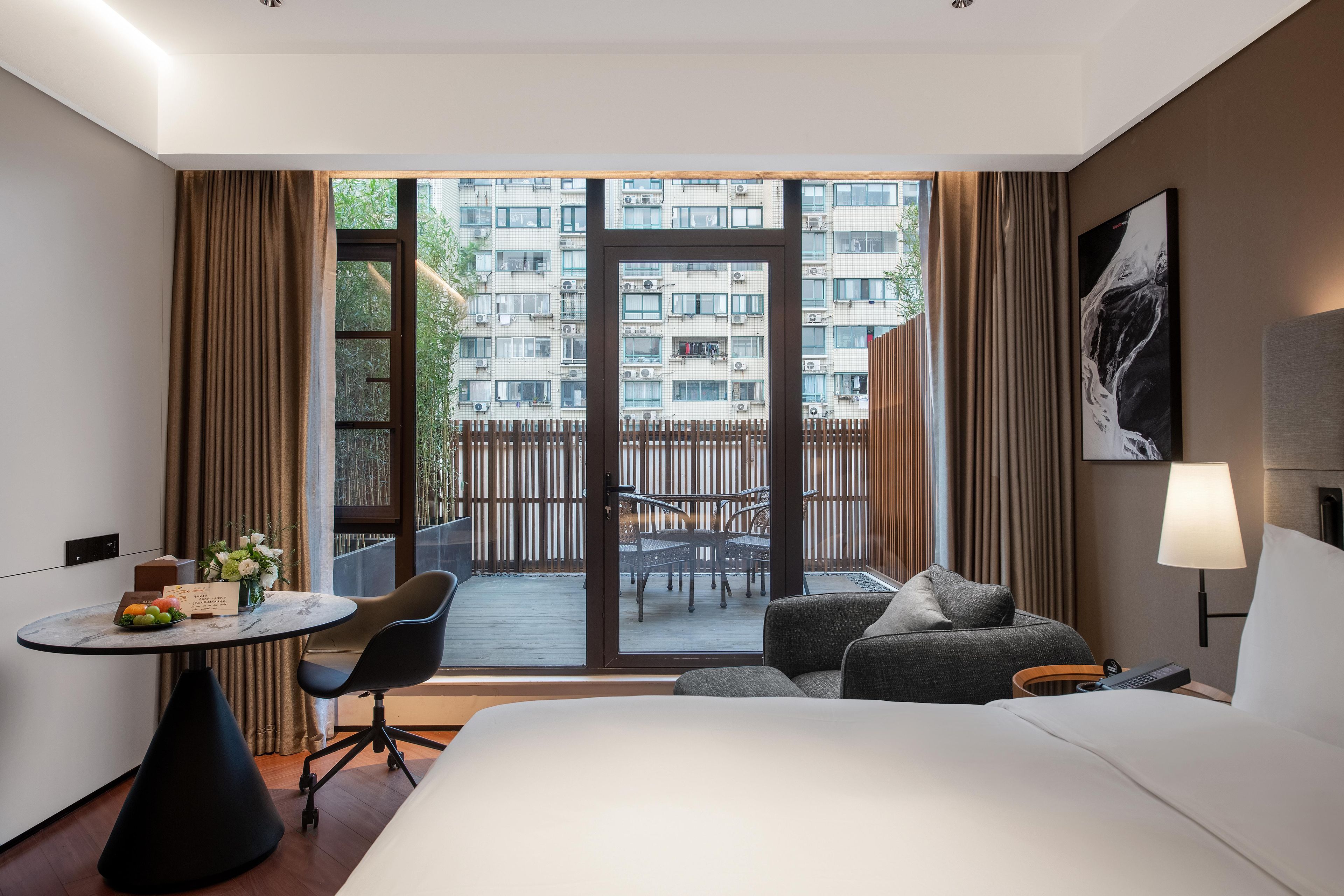 A hotel room with a bed, a desk with a chair, a small table with fruit and flowers, a sofa, and a view of an apartment building through a glass door.