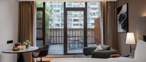 Une chambre d’hôtel avec un lit, un bureau et une chaise, une petite table avec des fruits et des fleurs, un canapé et une vue sur un immeuble d’habitation à travers une porte vitrée.