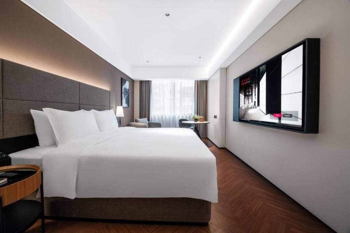 A modern hotel room with a large bed, a flat-screen TV mounted on the wall, and a view of the city through the window.