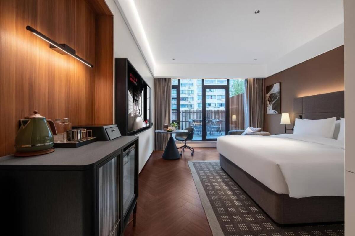 A modern hotel room with a bed, a desk with a kettle, a small table, and a view of the city through large windows.